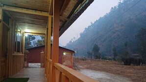 Un edificio de madera con un porche, un edificio rojo al fondo y una ladera boscosa.