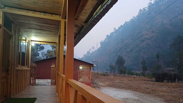 Un edificio de madera con un porche, un edificio rojo al fondo y una ladera boscosa.