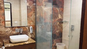 Un baño con pared de mármol, cabina de ducha con mampara de cristal, lavabo con encimera de granito y un inodoro.