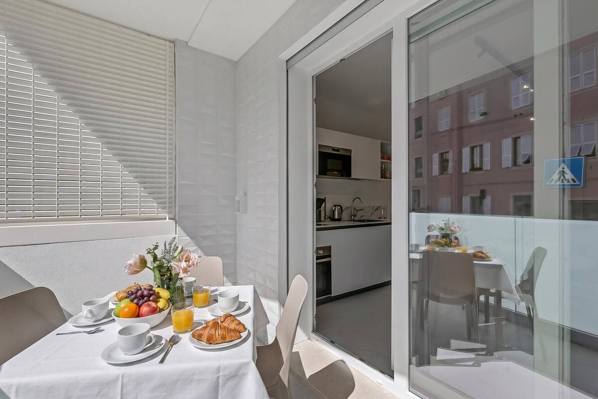 A balcony with a dining table set for breakfast, a kitchen in the background, and a view of a building.