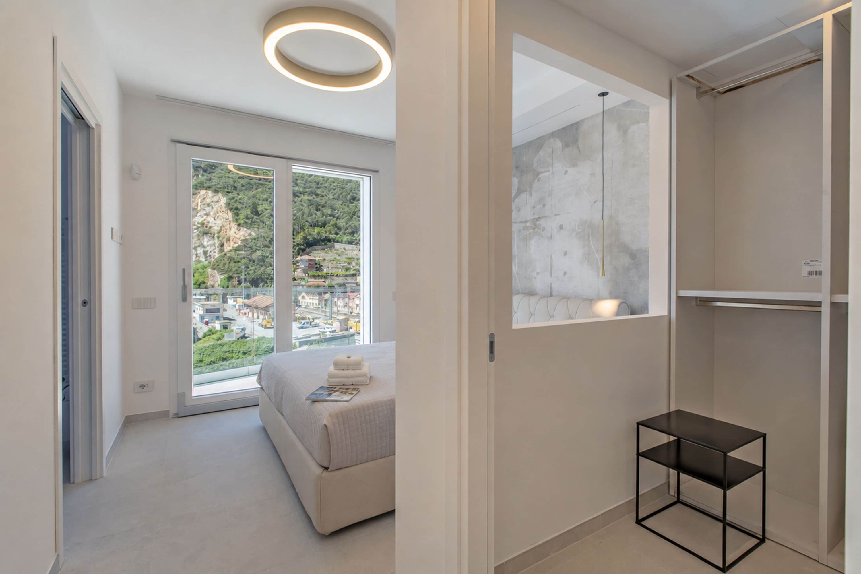 A modern bedroom with a large window, a bed, a bedside table, and a built-in wardrobe.