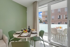 A dining area with a table set for two, a green couch, and a view of a building through a glass door. - Nua Boutique Apartaments (Finale Ligure)