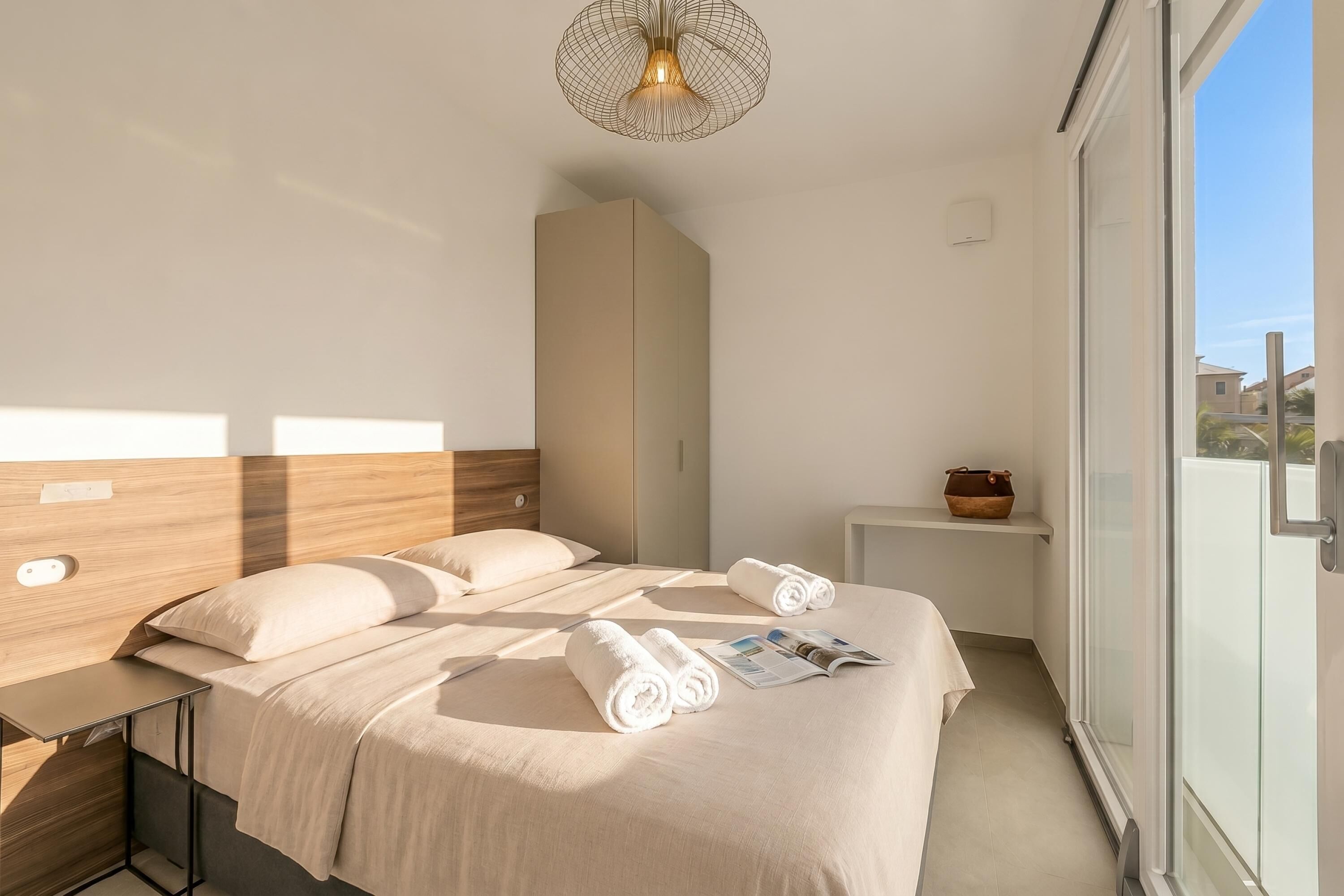 Une chambre d’hôtel moderne avec un grand lit, une table de chevet, une petite étagère et une vue sur l’extérieur, visible à travers une porte vitrée.