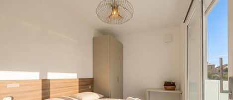 Une chambre d’hôtel moderne avec un grand lit, une table de chevet, une petite étagère et une vue sur l’extérieur, visible à travers une porte vitrée.