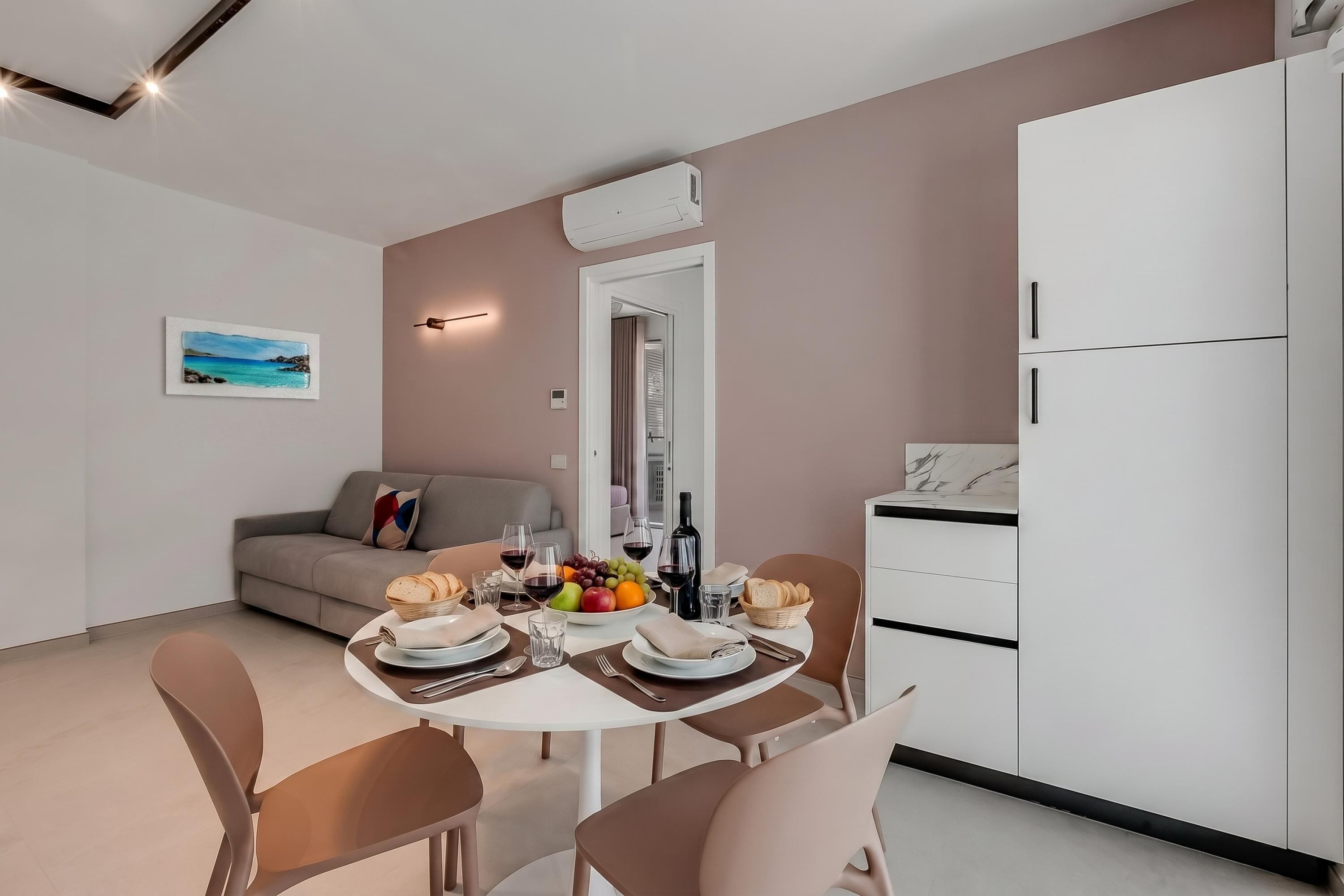 A modern living room with a dining table set for four, a sofa, a kitchen area with a refrigerator, and a painting on the wall.