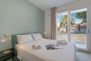 Habitación de hotel con cama, lámpara de noche, ventana con vista a palmeras y edificios, y una revista sobre la cama.