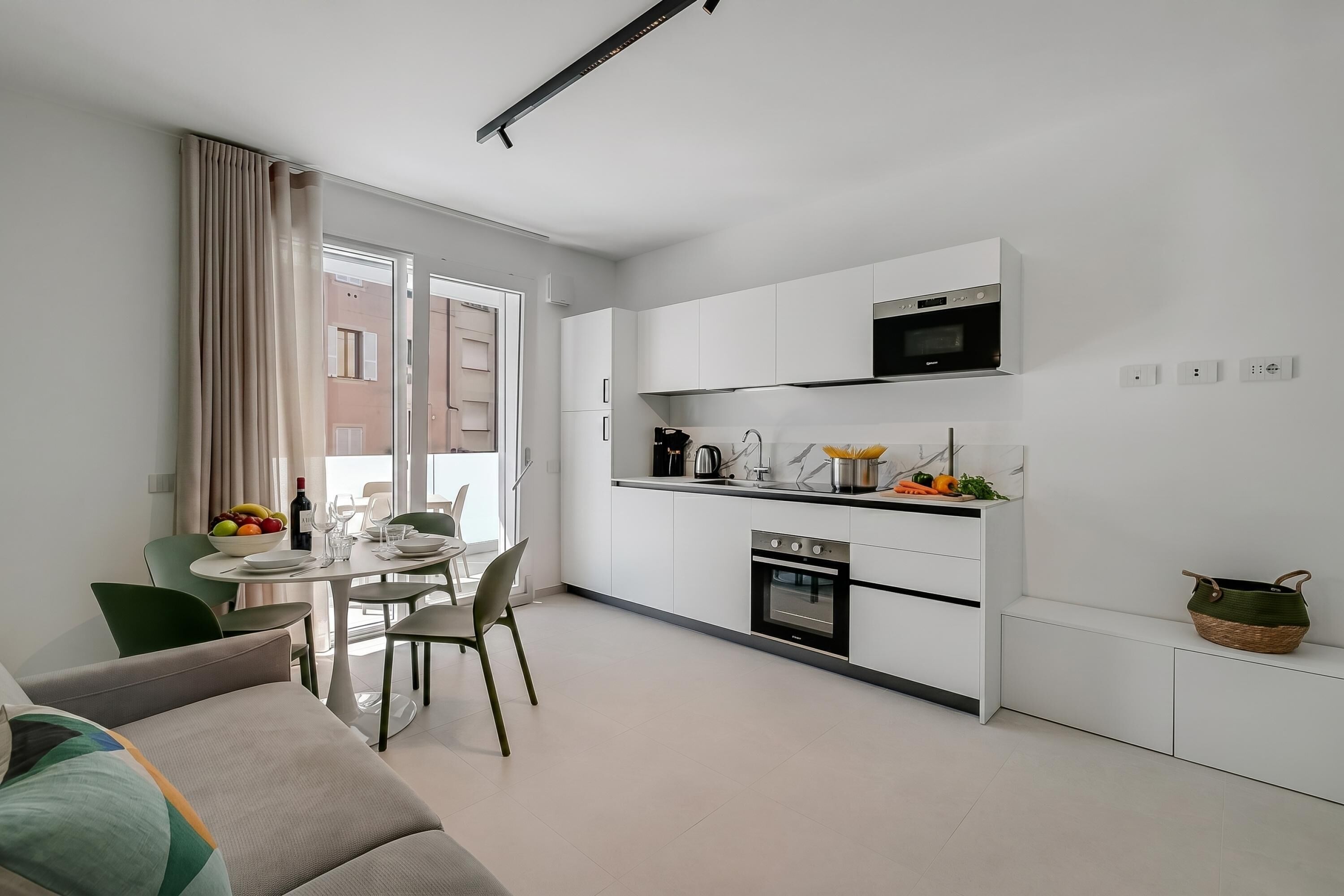A modern kitchen with white cabinets, a microwave, and an oven. There is a dining area with a table and chairs, and a sofa in the foreground.