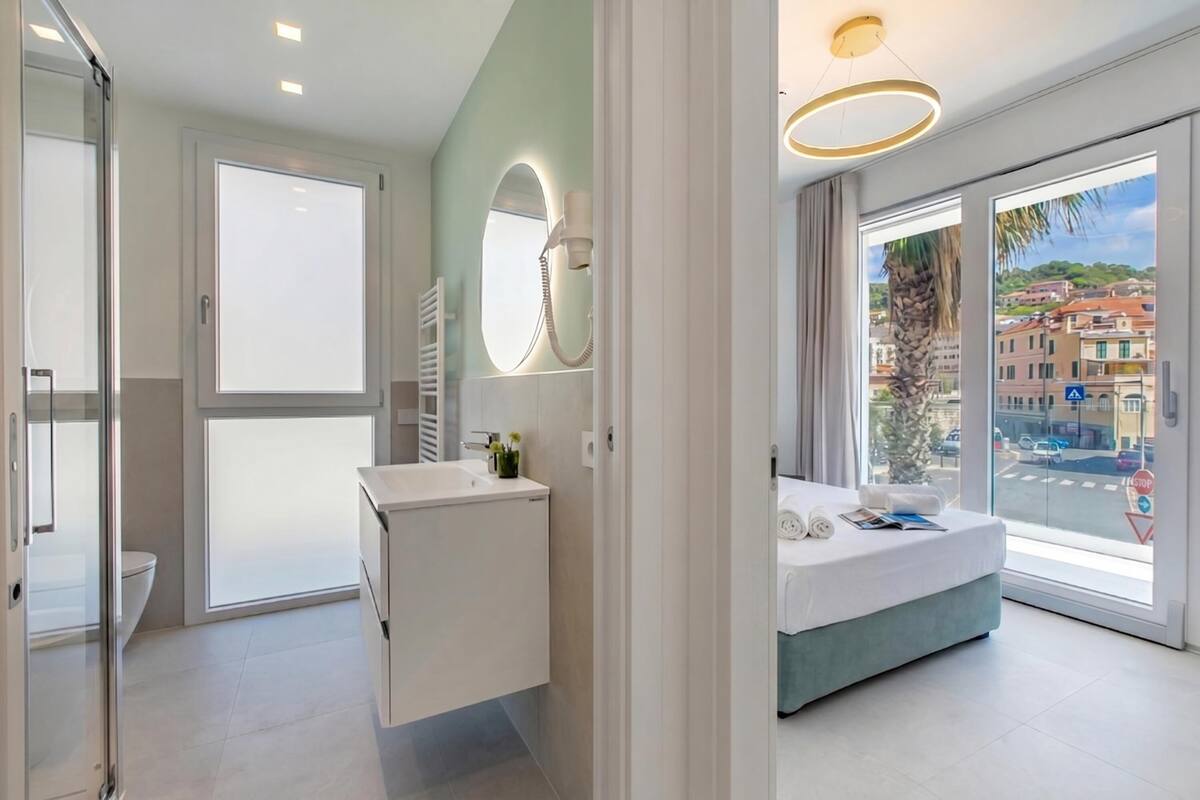 A modern hotel room with a large window, a bed, a bathroom with a mirror and sink, and a view of the city.