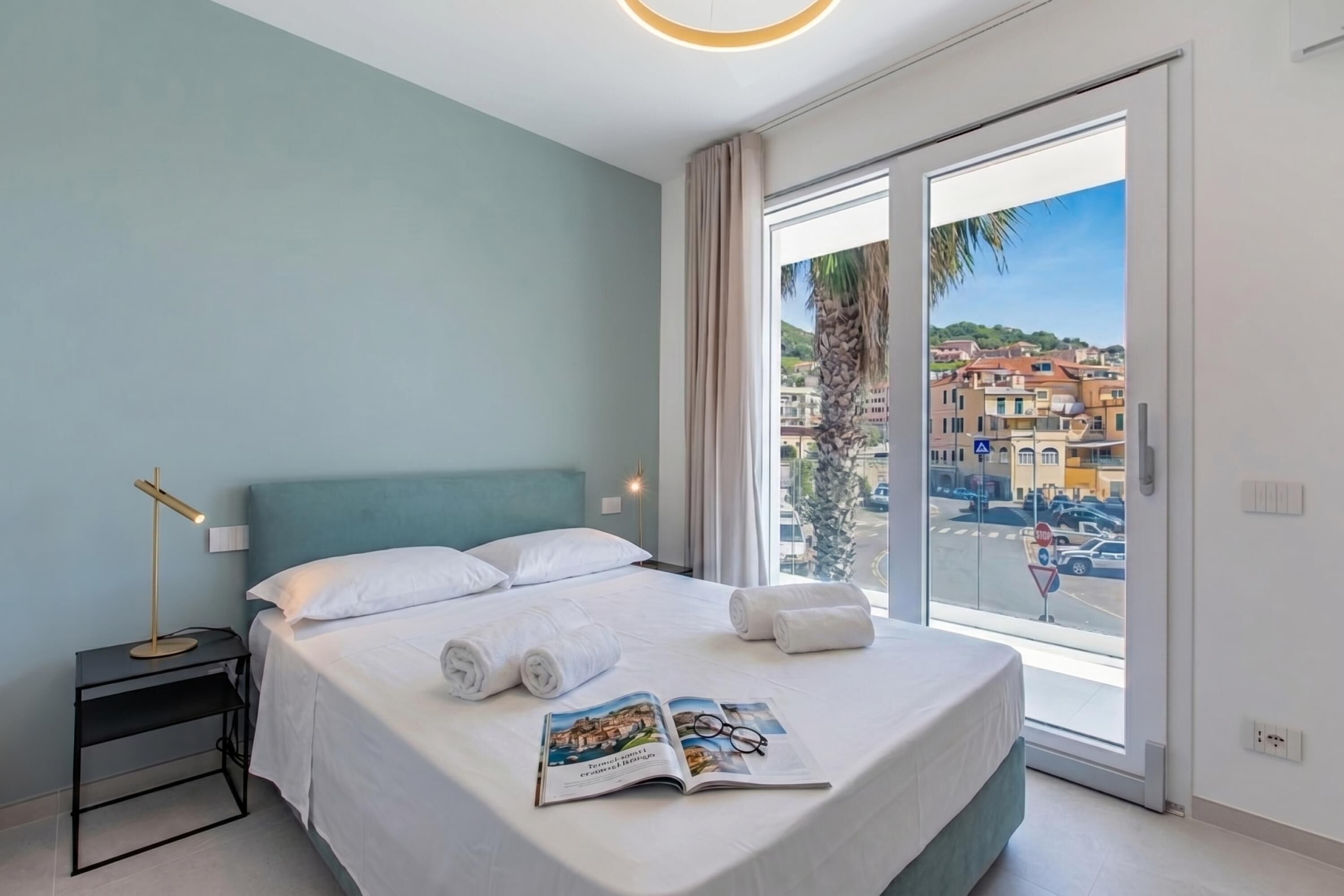 Ein Hotelzimmer mit Bett, Nachttisch, Leselampe, einem Fenster mit Blick auf Gebäude und eine Palme.
