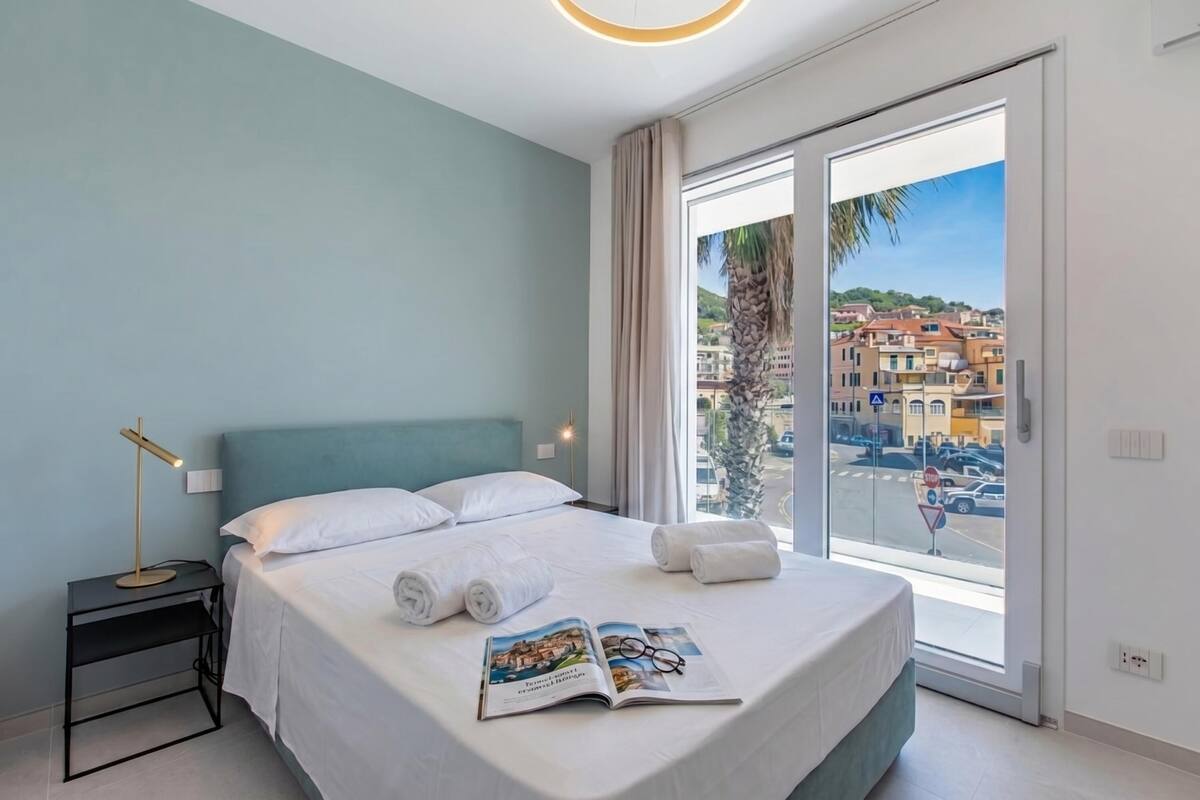 A hotel room with a bed, a nightstand, a reading lamp, a window with a view of buildings and a palm tree.