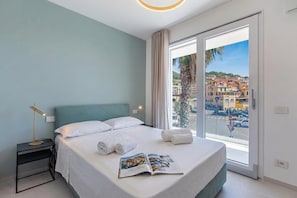 Habitación de hotel con cama, mesita de noche, lámpara de lectura, ventana con vistas a edificios y una palmera.