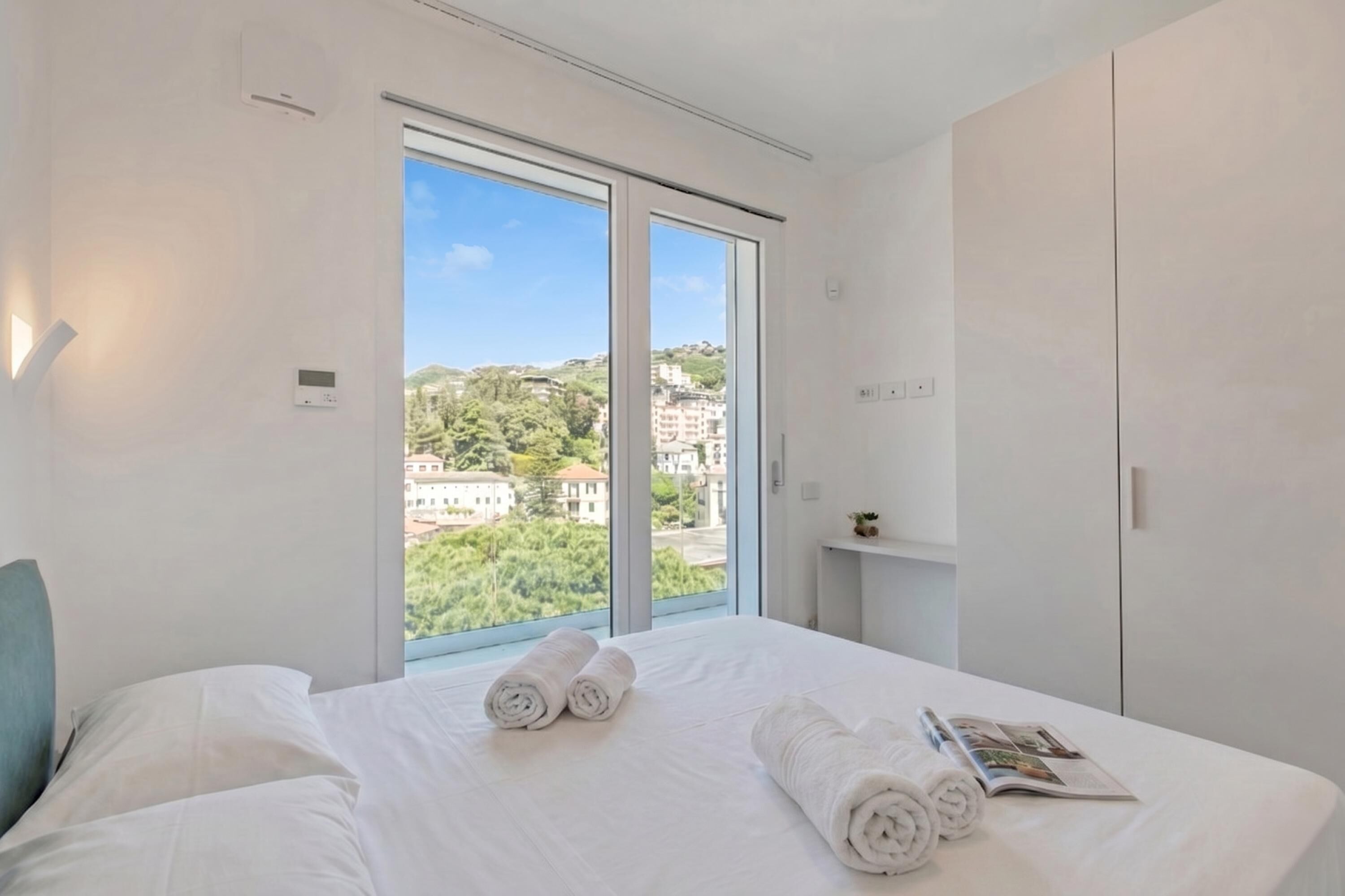 Habitación de hotel con un ventanal grande, una cama con ropa de cama blanca y vistas a edificios y zonas verdes.