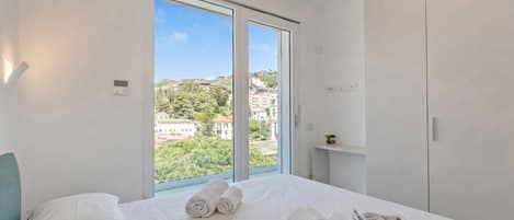 A hotel room with a large window, a bed with white linens, and a view of buildings and greenery.