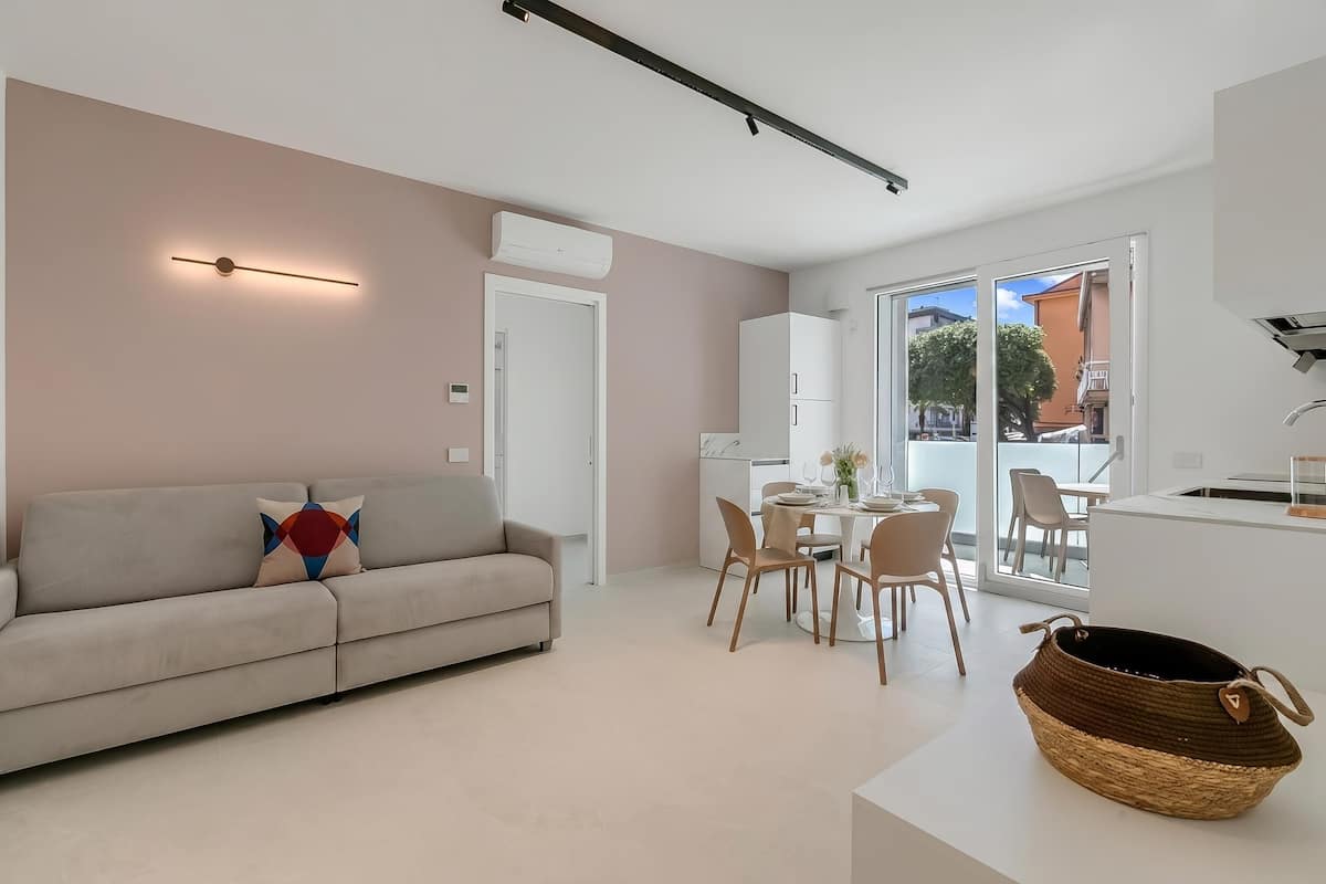 A modern living room with a sofa, a dining area with chairs, a kitchen area, and a large window.