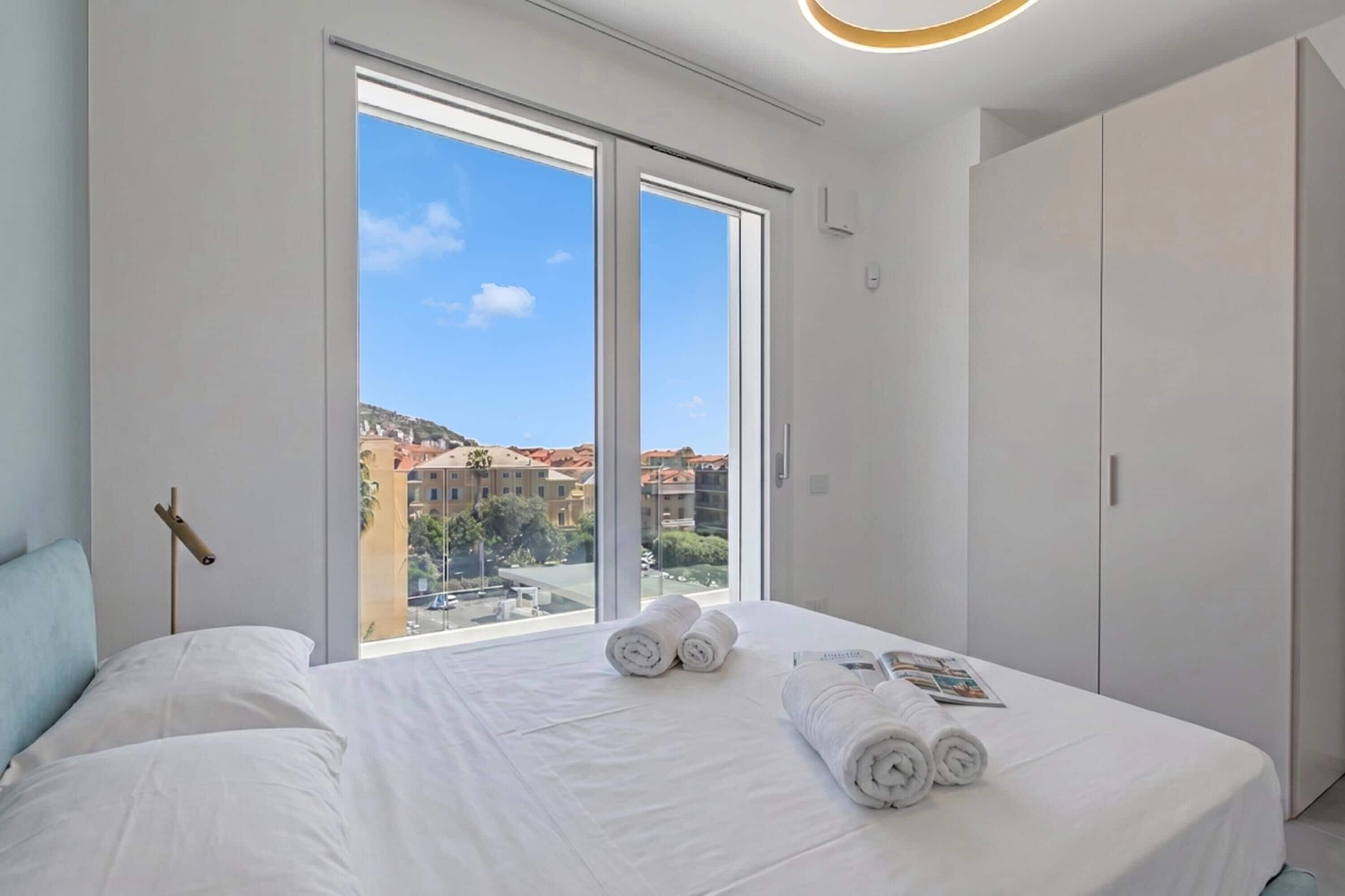 Une chambre d’hôtel avec une grande fenêtre, un lit avec du linge de maison blanc, une armoire et une table de chevet.