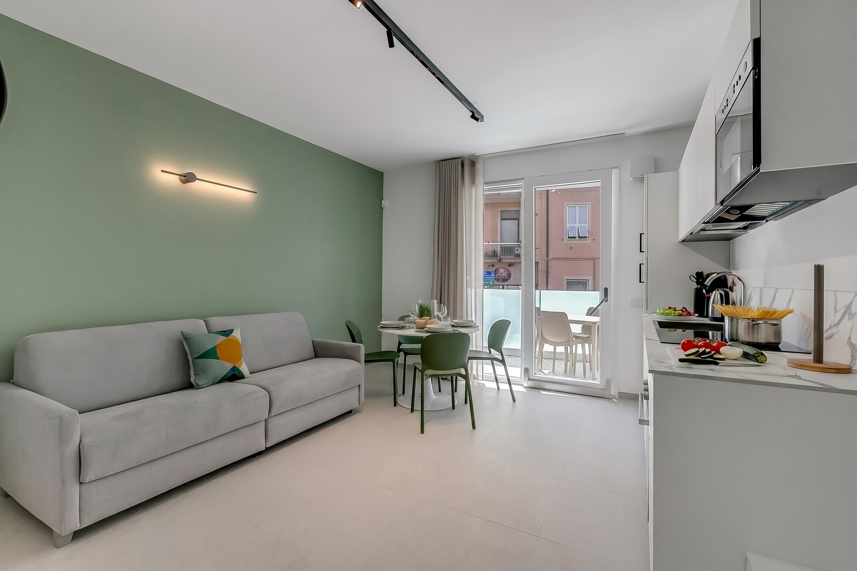 Ein modernes Wohnzimmer mit einer Couch, einem Esstisch und Stühlen, eine Küche mit Herd und Arbeitsplatte sowie ein Blick auf ein Gebäude durch eine Glastür.