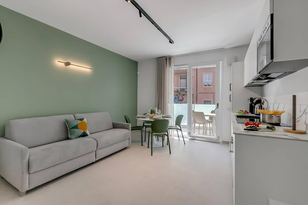 A modern living room with a sofa, dining table, and chairs, a kitchen with a stove and counter, and a view of a building through a glass door.