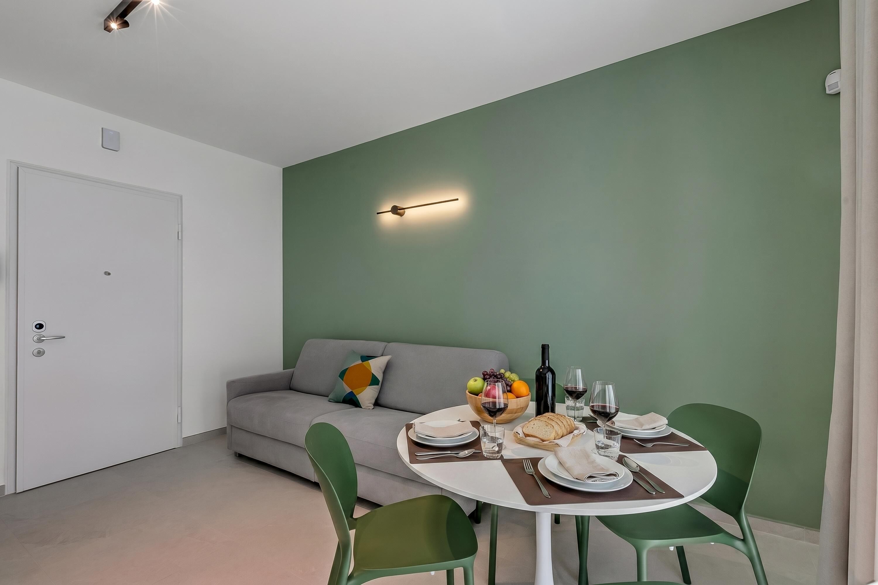 A modern room with a dining table set for two, a sofa, and a green wall.