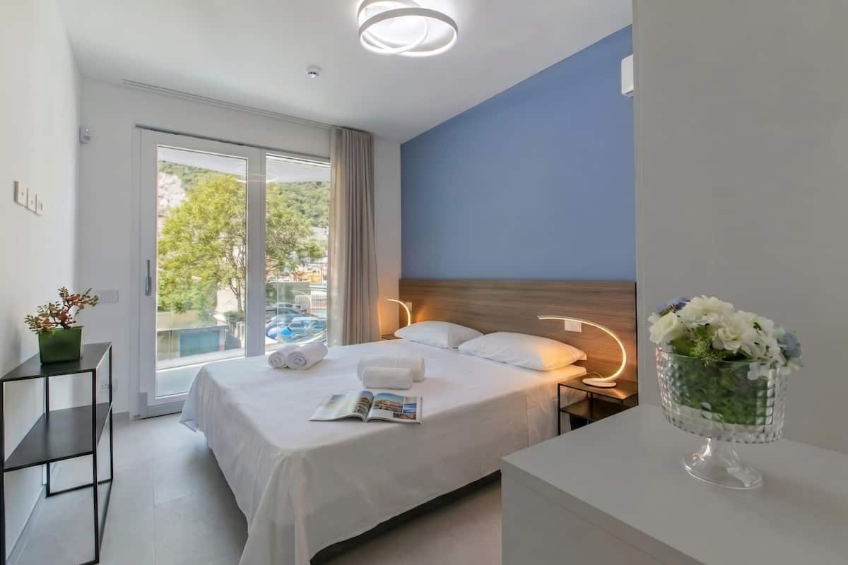 A hotel room with a bed, a bedside table, a nightstand, a window with a view, and a vase of flowers.