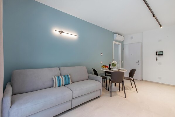 Una sala de estar moderna con un sofá, una mesa de comedor con sillas y una lámpara fijada a la pared.