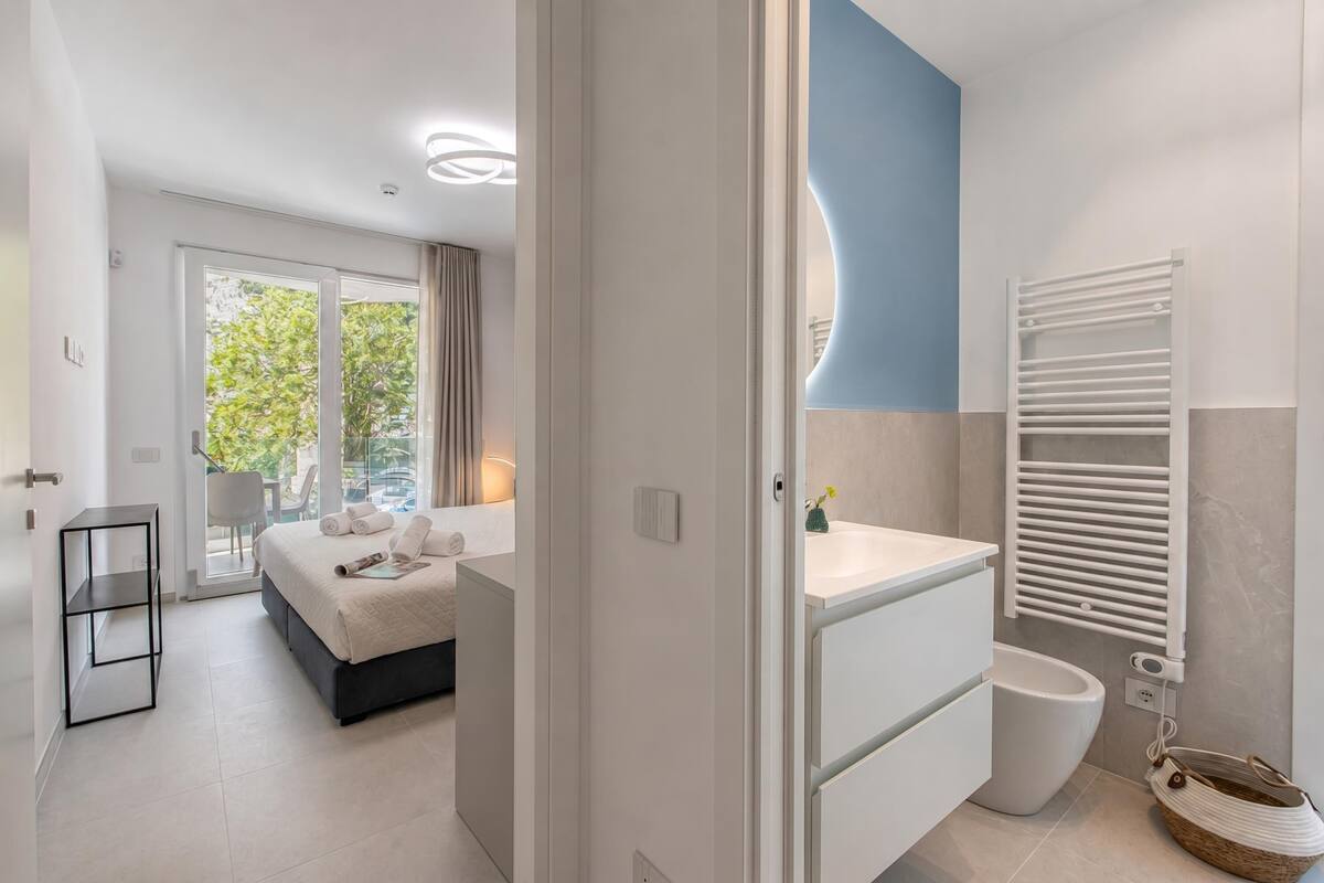 A modern hotel room with a bed, a bathroom with a white sink and toilet, and a balcony with a view of greenery.
