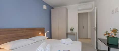 A hotel room with a bed, a bedside table, a potted plant, and a door leading to another room.