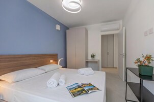 Una camera d'albergo con un letto, un comodino, una pianta in vaso e una porta che dà su un'altra stanza.