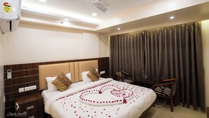 A hotel room with a bed, a ceiling fan, a window with curtains, a bedside table, and a chair. - HOTEL M K GRAND ISHWAR TOWER (Varanasi)