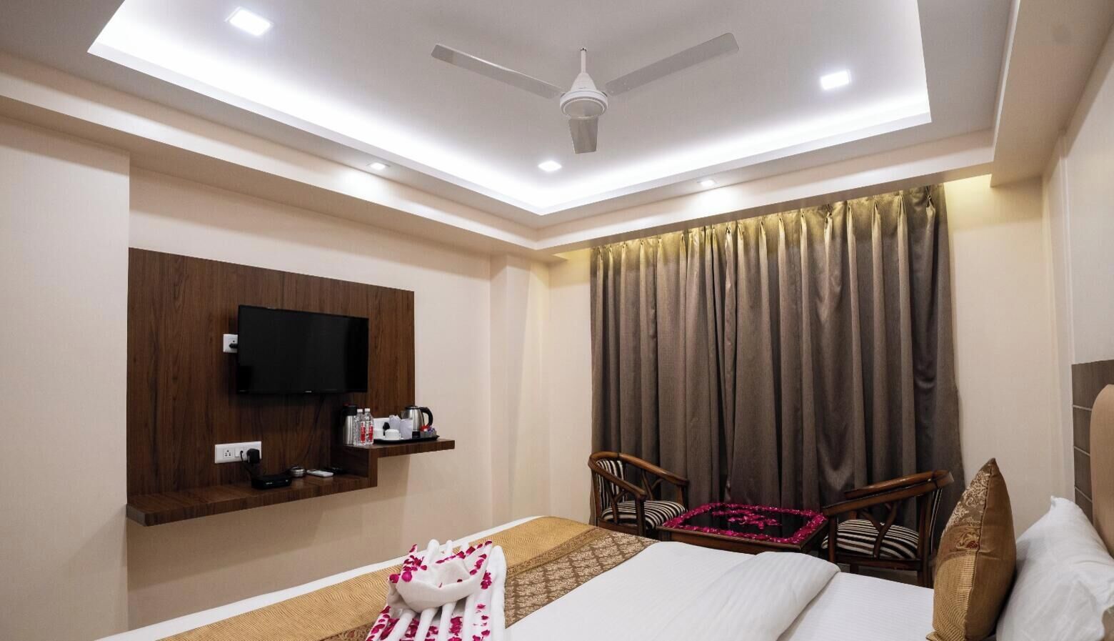 A hotel room with a bed, a TV mounted on the wall, a ceiling fan, and a window with curtains.