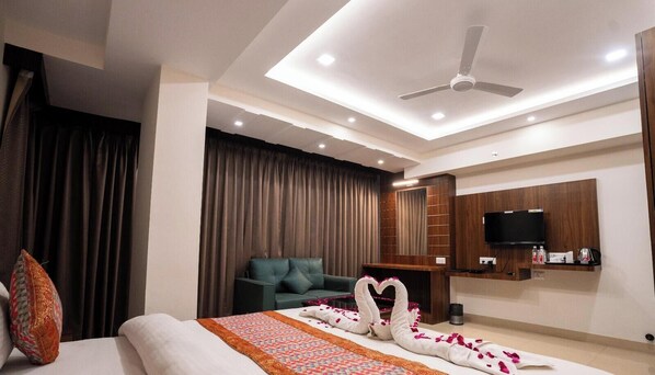 A hotel room with a bed, a green sofa, a TV mounted on the wall, and a ceiling fan. - HOTEL M K GRAND ISHWAR TOWER (Varanasi)