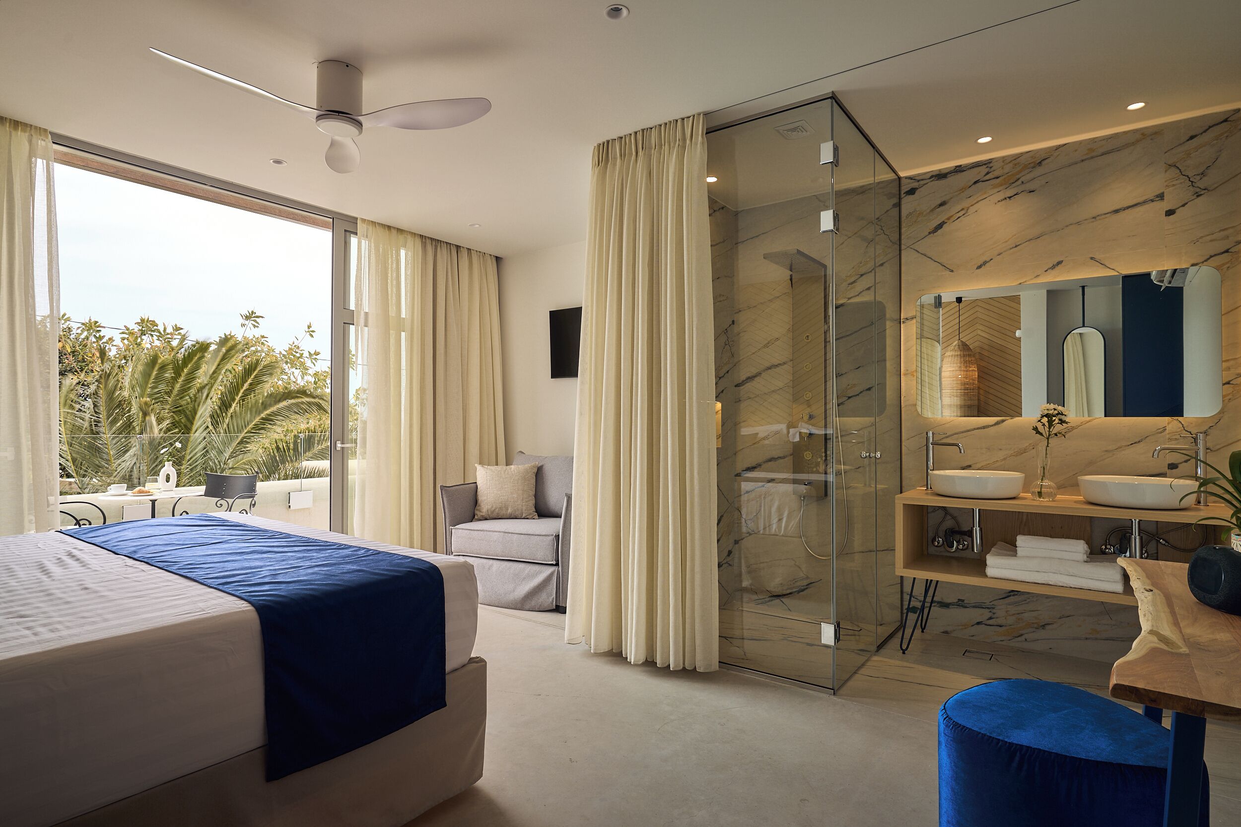 A modern hotel room with a large bed, a seating area with a sofa and armchair, a glass-enclosed shower, and a marble bathroom.