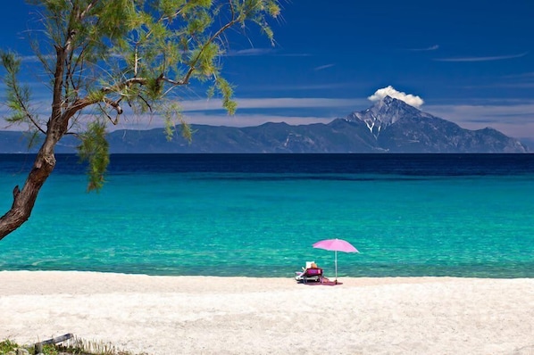 A beach with clear turquoise water, a mountain in the background, and a person under a pink umbrella.