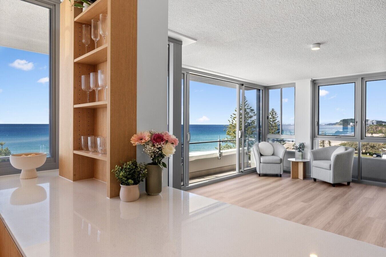 A modern living room with a large window overlooking the sea, a wooden bookshelf, and a seating area with two armchairs.
