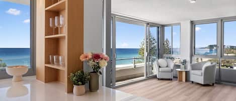 A modern living room with a large window overlooking the sea, a wooden bookshelf, and a seating area with two armchairs.