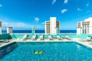 Ein Pool auf dem Dach mit klarem, blauem Wasser, umgeben von Liegestühlen und Blick auf das Meer und die Gebäude.