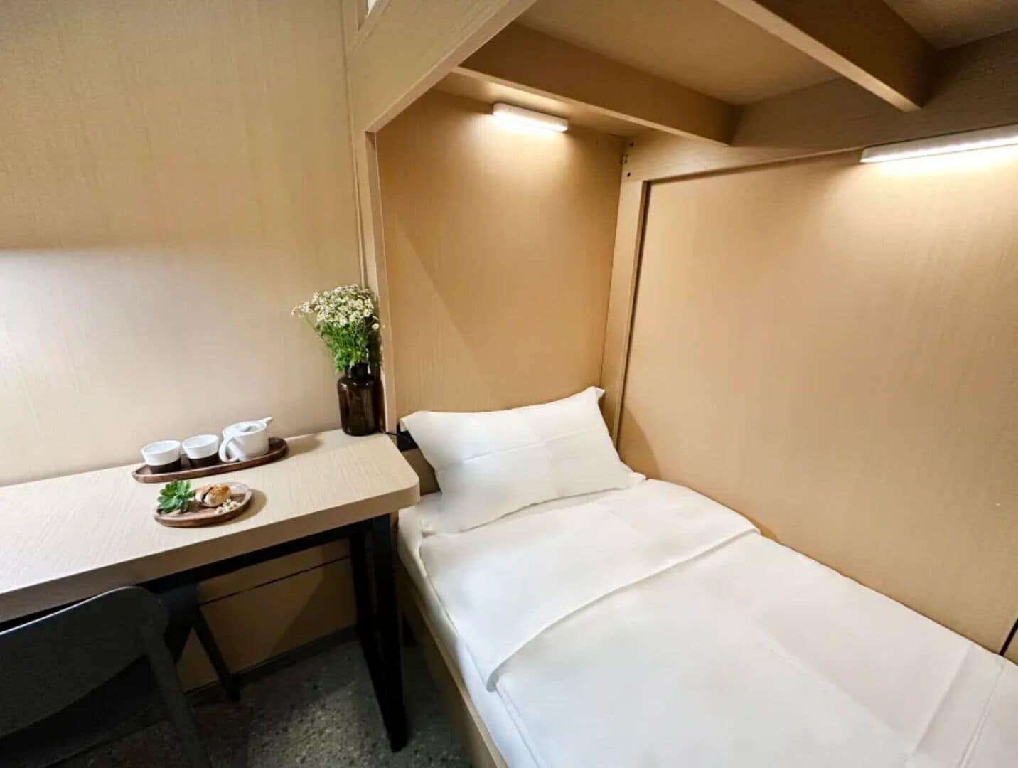 Une petite pièce comprenant un lit, une petite table avec des tasses et une assiette, et un vase avec des fleurs.