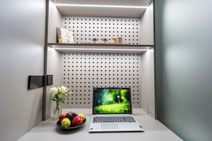 A laptop on a desk with a perforated wall behind it, a vase with flowers, and a bowl of fruit.