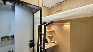 A compact hotel room with a bunk bed, a desk with a laptop, a small seating area, and a minibar.