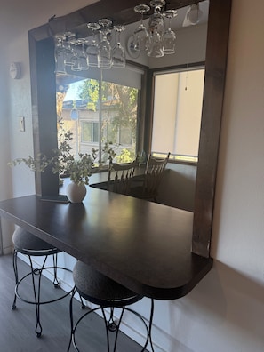 Un comedor con una mesa de madera oscura, dos sillas, una planta en maceta y una ventana con vistas a edificios y árboles.