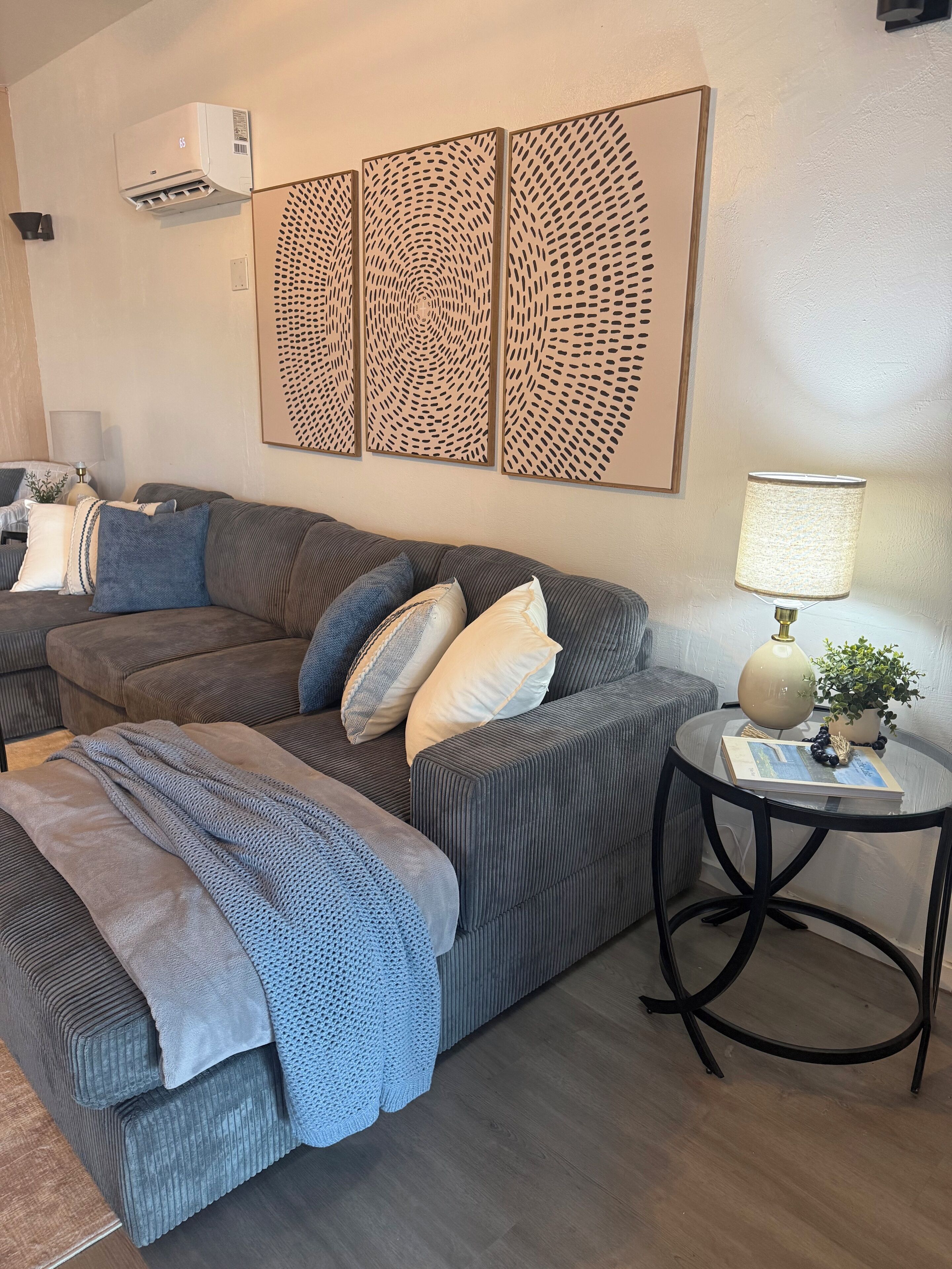 A living room with a grey sofa, a round side table with a lamp, and a wall-mounted air conditioner.