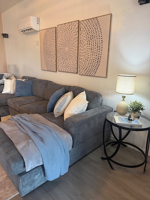 Un salón con un sofá gris, una mesita redonda con una lámpara y un aire acondicionado instalado en la pared.