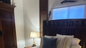 A neatly made bed with multiple pillows and a blanket, a wooden wardrobe, a nightstand with a lamp, and a wall-mounted air conditioning unit.