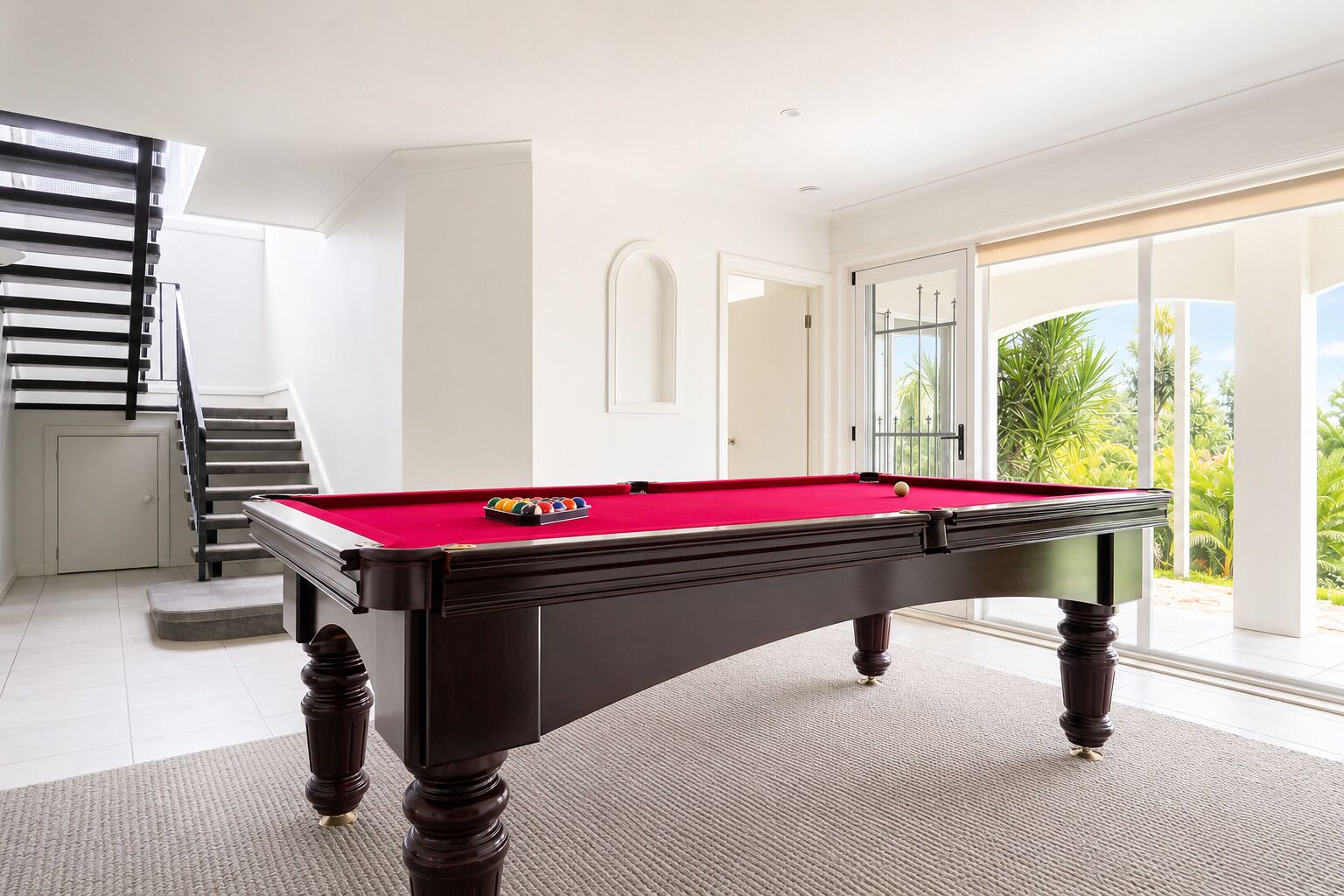 A pool table in a room with a staircase, large windows, and a view of greenery.