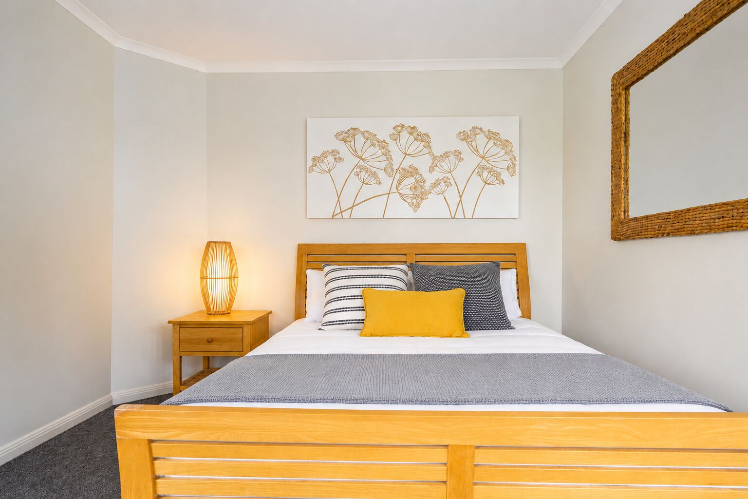 Un dormitorio con una cama de madera, una mesita de noche con una lámpara, un espejo en la pared y una obra de arte floral.