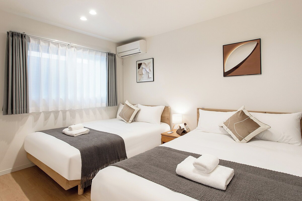 A hotel room with two beds, a window with curtains, an air conditioning unit, a bedside table with a lamp, and artwork on the walls.
