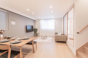 A modern living room with a sofa, a dining table set with plates, a TV mounted on the wall, and a staircase leading to another level. - SHOTENSHITA (Osaka)