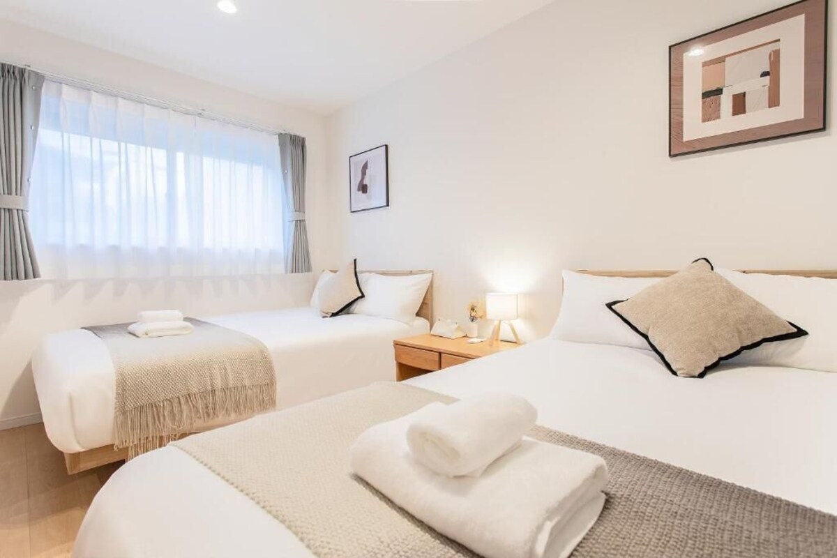 A hotel room with two beds, a bedside table with a lamp, a window with curtains, and a framed picture on the wall.