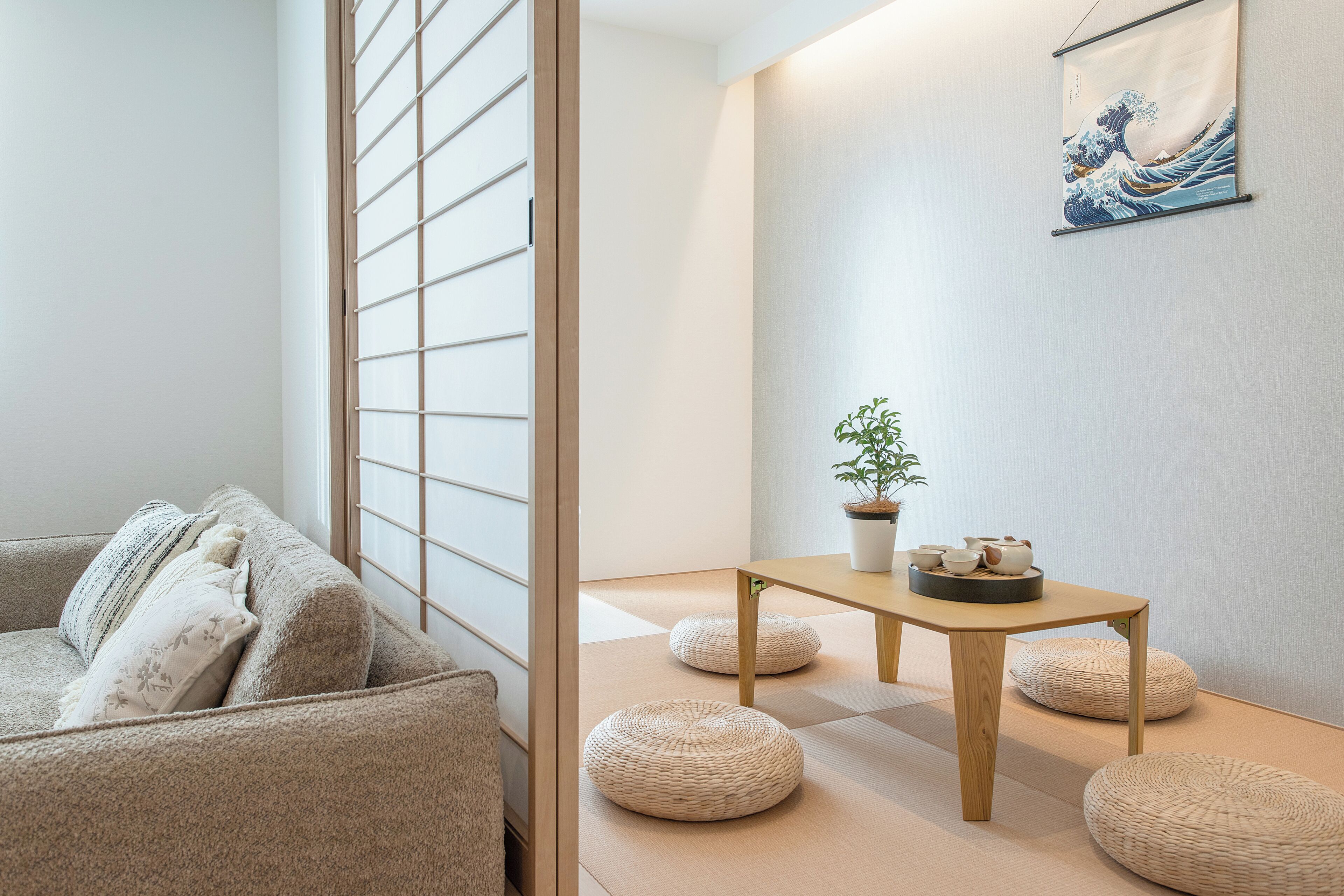 A room with tatami flooring, a low wooden table with a plant and teaware, a sofa with cushions, and a shoji screen.