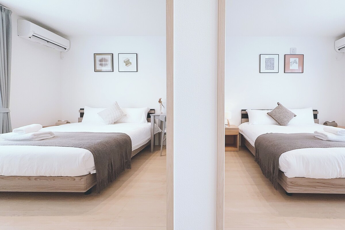 A mirrored hotel room with two beds, white bedding, a small bedside table, and wall-mounted air conditioning units.