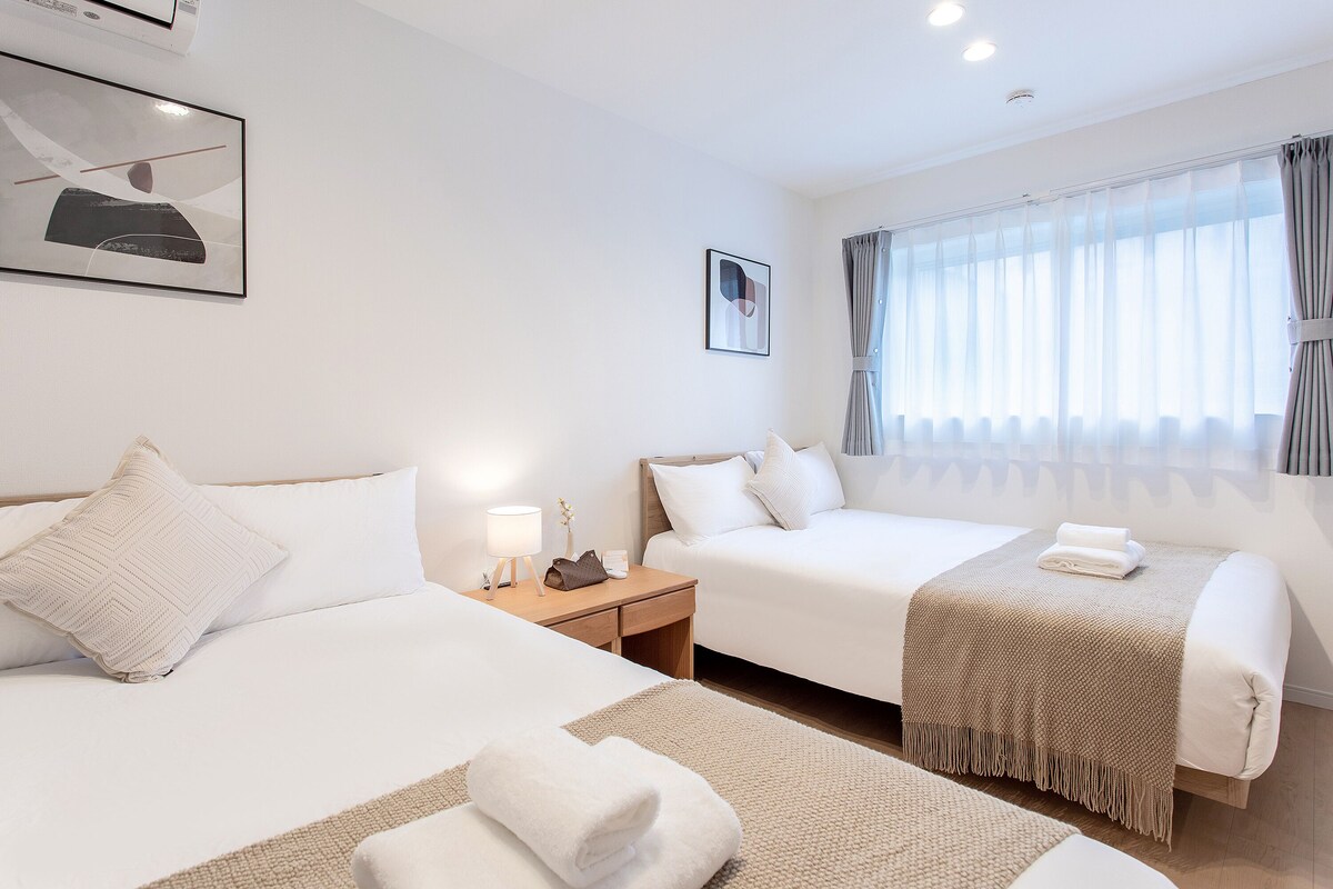A hotel room with two beds, a nightstand with a lamp, a window with curtains, and artwork on the walls.
